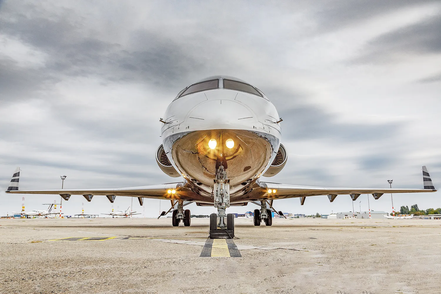 Front view of a private jet on the tarmac with its landing lights on and an overcast sky in the background.