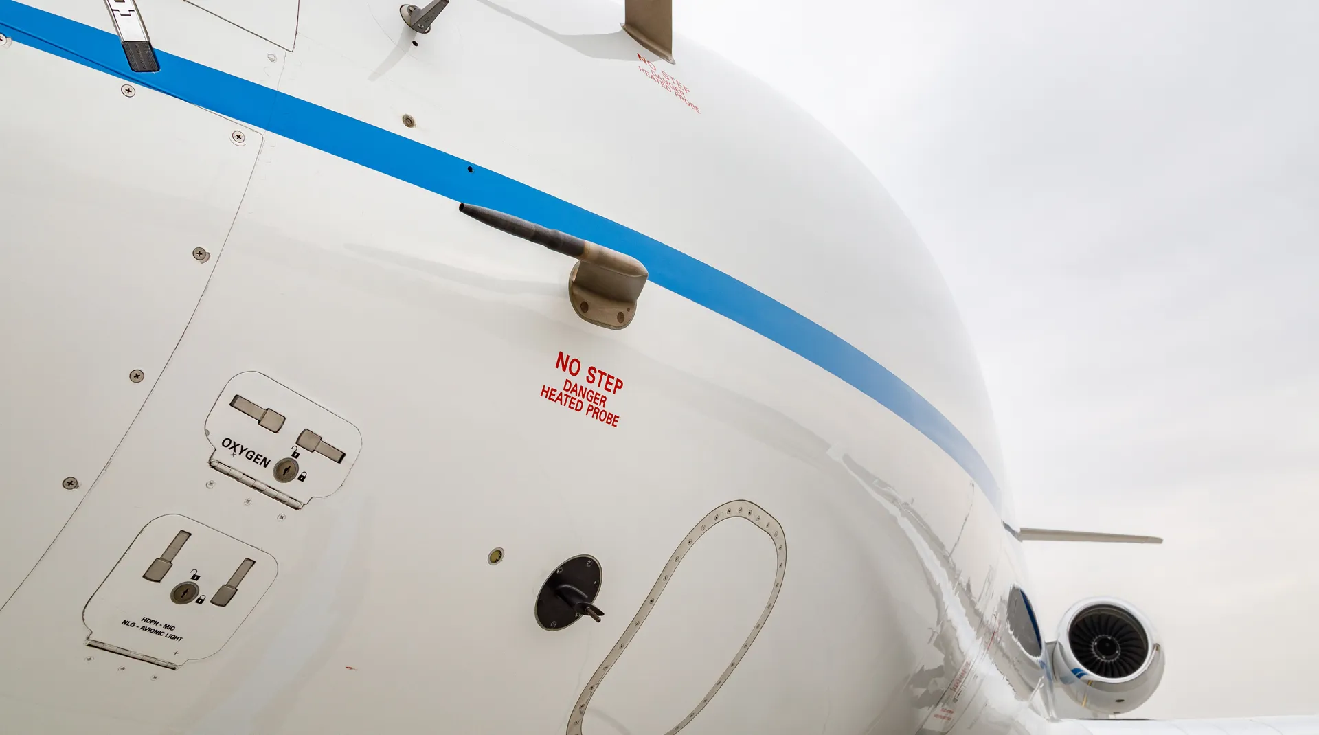 Close-up of a white private jet’s nose featuring a blue stripe, visible safety labels, and a subtle overcast sky in the background.